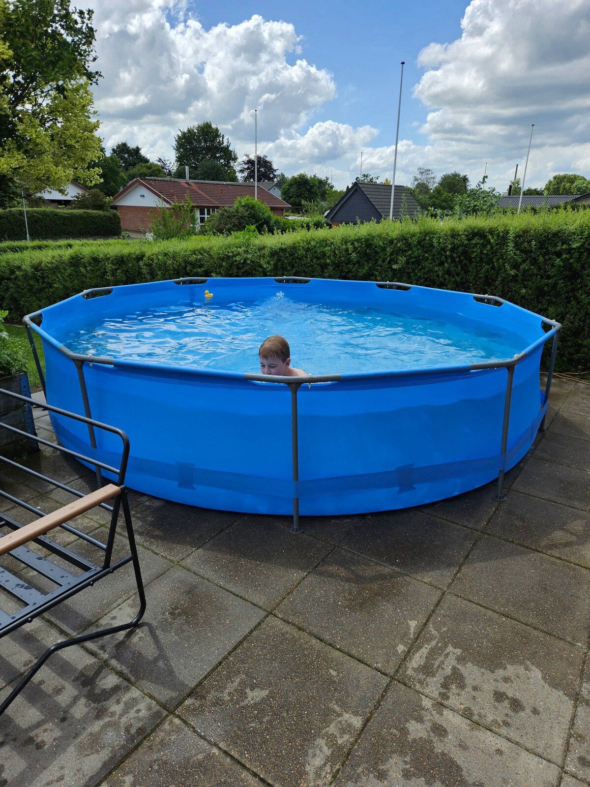 A round above-ground pool is situated outdoors, surrounded by lush greenery and low hedges. Sunlight reflects off the clear water, creating a bright and inviting atmosphere. Patrons can enjoy the privacy offered by the surrounding landscape while relaxing on the nearby paved area with seating.