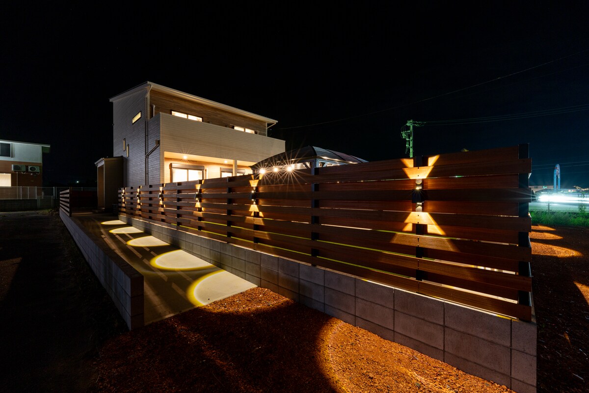 The exterior of a newly constructed building is illuminated at night, featuring a modern wooden fence that lines the pathway. Soft lighting highlights the entrance, creating a welcoming ambiance. The clean lines of the architecture are complemented by a gravel surface, enhancing the overall contemporary feel.
