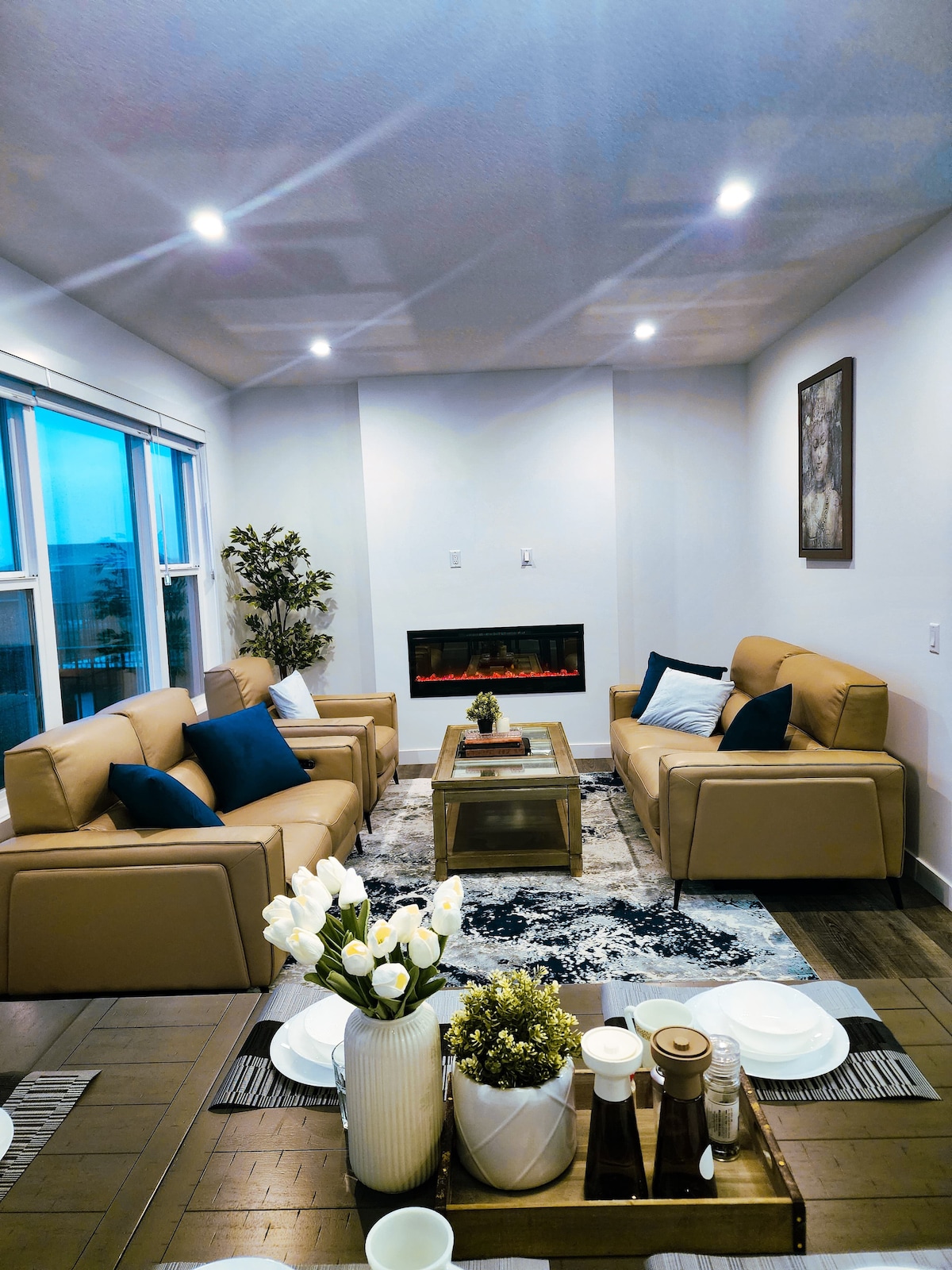 A modern living area showcases two tan sofas with blue pillows, arranged around a wooden coffee table. A light area rug lies beneath, complementing the neutral color scheme. Near the window, a potted plant adds a natural touch, while a wall-mounted electric fireplace creates a cozy focal point.