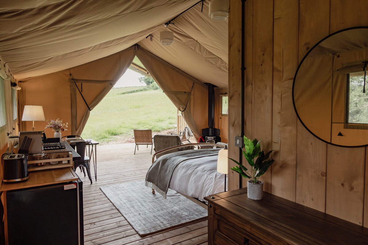 The interior of a canvas lodge is beautifully arranged with wooden walls and a cozy atmosphere. A spacious bed is complemented by bedside lighting. A small dining table and a comfortable chair are positioned near a window, providing views of the surrounding countryside.