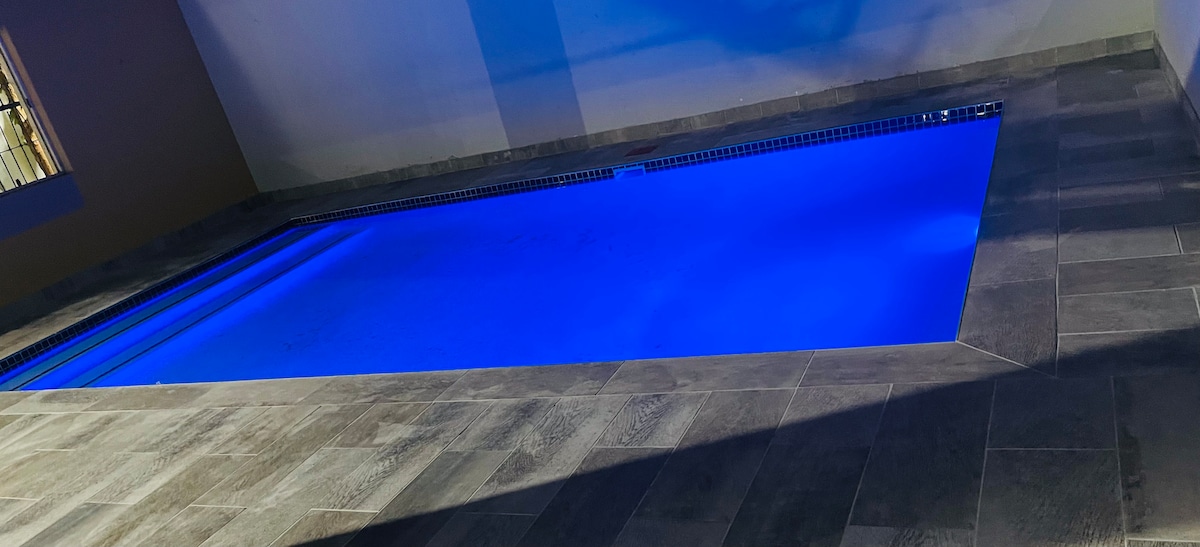 A sleek indoor pool is illuminated with a calming blue light, set against a modern tiled surface. The tranquil water reflects the lighting, creating a serene ambiance. The space is designed for relaxation and refreshment, enhancing the indoor experience.