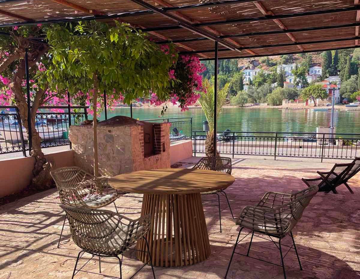 An outdoor seating area is furnished with a round wooden table surrounded by four woven chairs. A stone barbecue grill is positioned nearby. A pergola offers shade, adorned with vibrant pink bougainvillea and green foliage. The tranquil water reflects the surrounding landscape.
