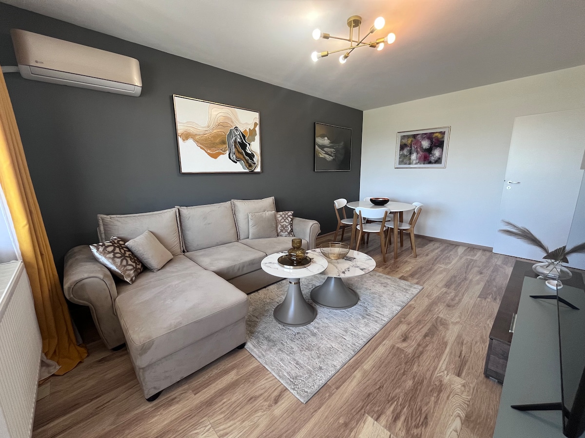 A bright living area is furnished with a light-colored sectional sofa and two marble-topped tables. A dining set for four is positioned nearby. Walls feature modern artwork, and a window with sheer curtains allows natural light to brighten the space.