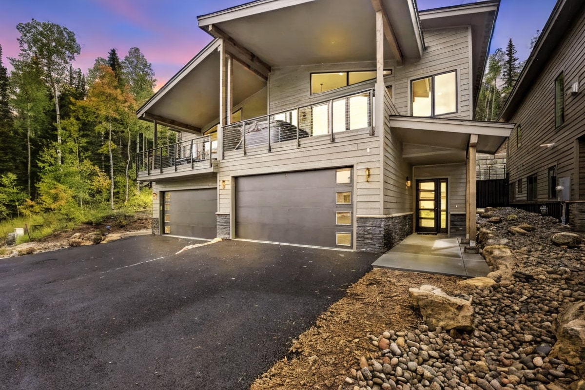 A modern mountain home is surrounded by tall trees, featuring a spacious driveway and a two-car garage. Large windows create a bright, open feel, and the entrance is framed by rustic stone accents, showcasing a blend of contemporary design and natural beauty.