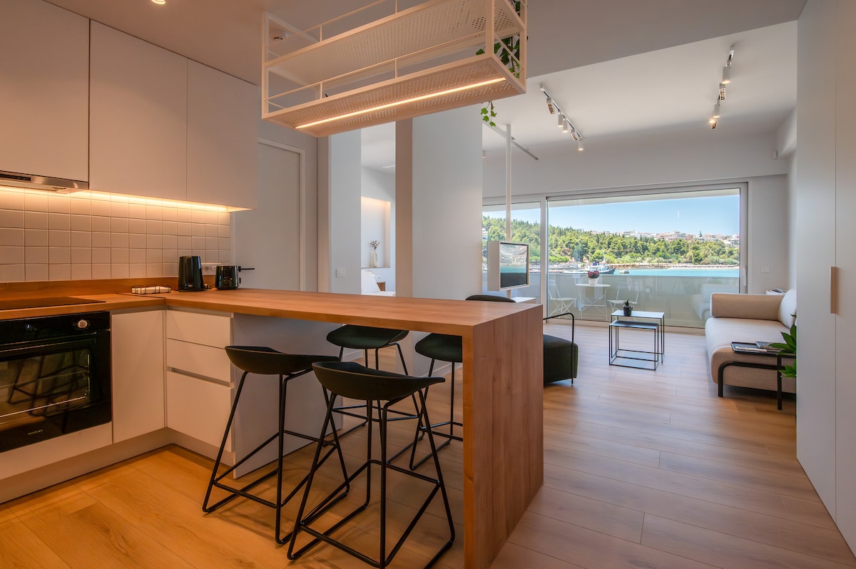 A modern kitchen space features a wooden island with three black stools. Sunlight highlights the bright cabinetry and clean countertops. The adjoining living area is partially visible, showcasing large windows with views of greenery. Light-colored flooring enhances the open, airy atmosphere.
