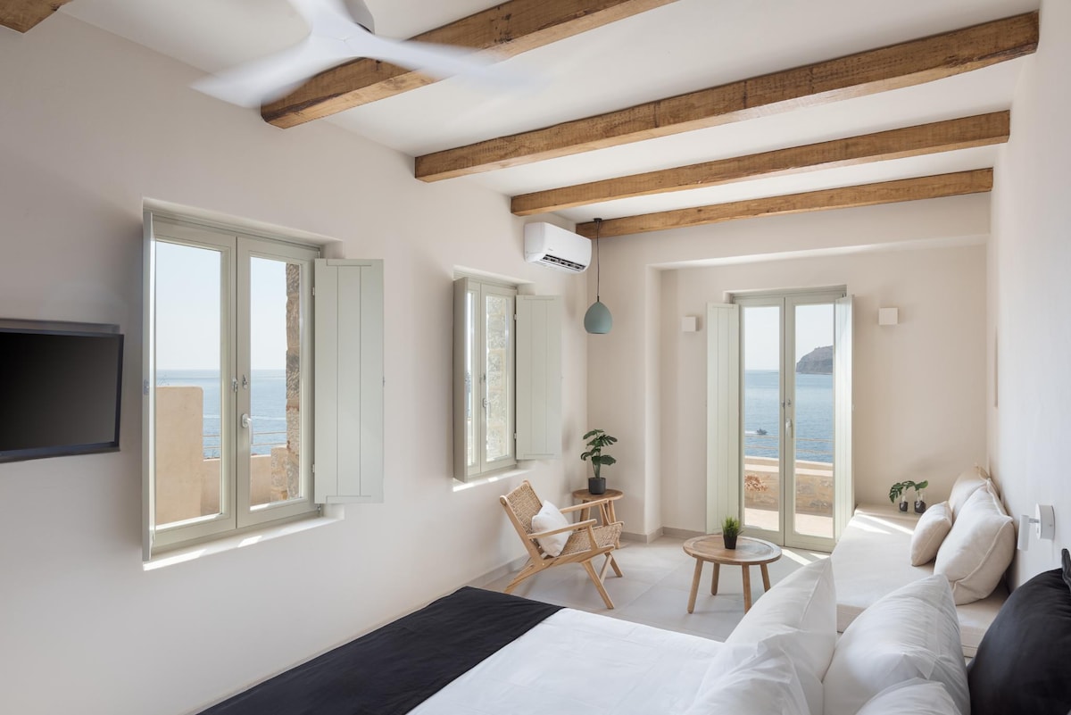 A bright living space features multiple windows inviting natural light and showcasing a scenic view of the sea. A cozy seating area includes a light-colored chair and a round table, while exposed wooden beams add to the room's airy ambiance.