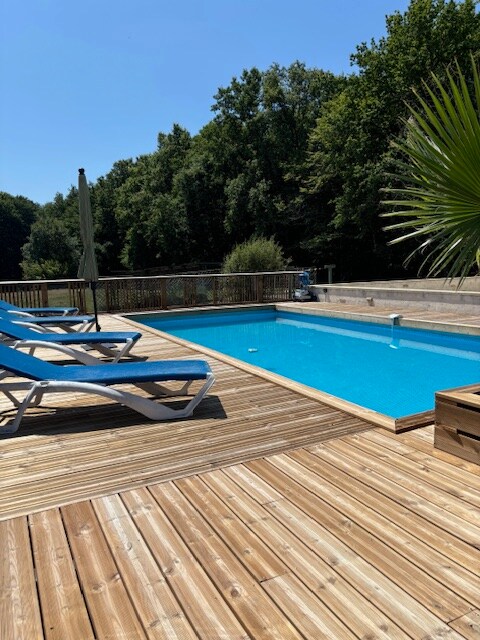 A wooden deck surrounds a clear blue swimming pool, inviting relaxation. Lounge chairs in a light blue color are positioned for comfort. Lush greenery is visible in the background, enhancing the natural setting and offering a peaceful ambiance.