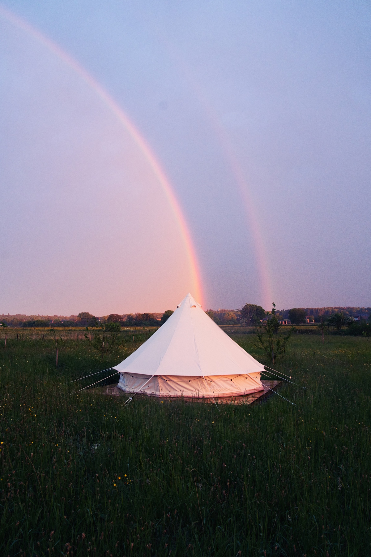 A spacious white tent is situated in a grassy field, framed by a vibrant rainbow arching across the sky. The surrounding area features tall grasses and scattered trees, with soft hues of sunset lighting the scene.
