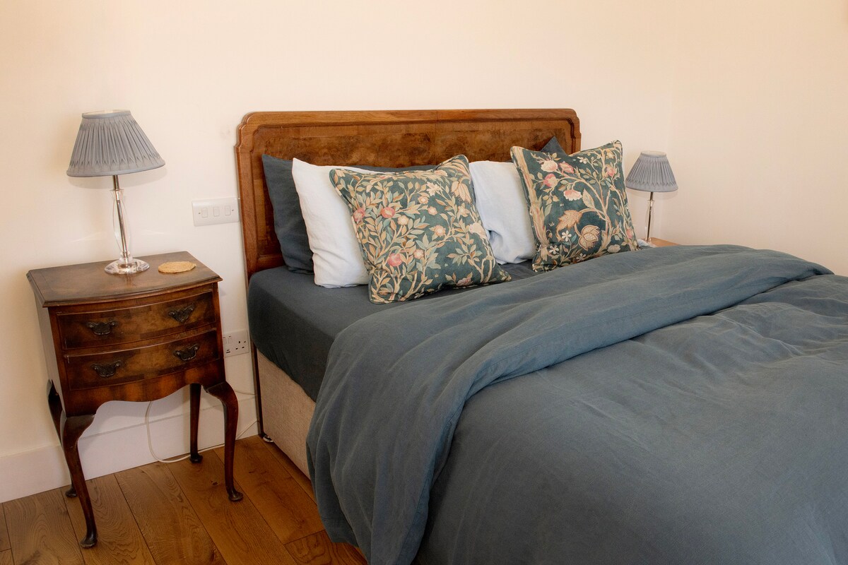 A cozy double bed is framed by soft, decorative pillows featuring floral patterns. Two small bedside lamps cast a gentle light. A wooden bedside table, adorned with a coiled placemat, stands next to the bed, blending seamlessly with the warm wooden flooring.