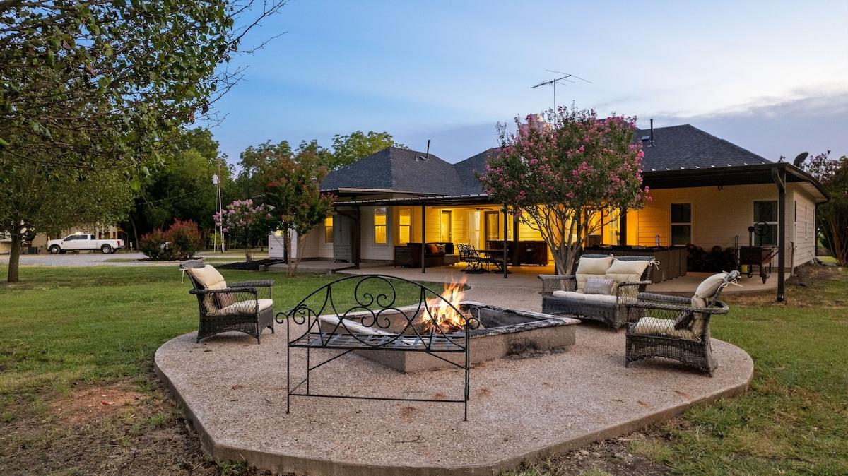 An outdoor fire pit area features a stone base surrounded by comfortable seating. Two stylish chairs and a sofa provide a casual gathering space, with a warm fire burning in the center. Soft lighting from the nearby house creates a cozy ambiance as dusk falls.