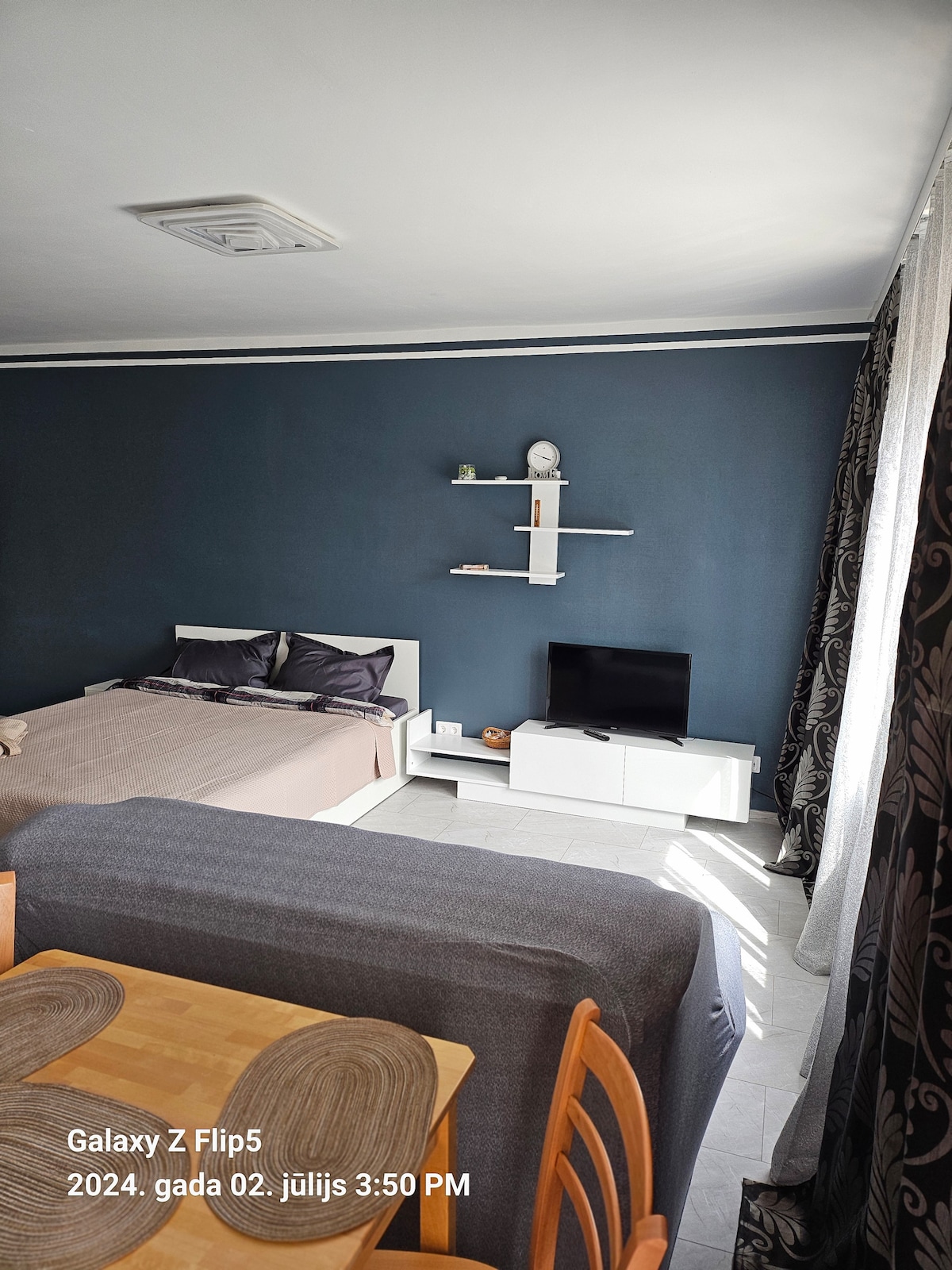 A studio apartment features a spacious bed with neutral bedding set against a dark blue wall. A small television is placed on a white media unit. A dining area with wooden chairs and a table is positioned near a sofa, providing a comfortable living space.