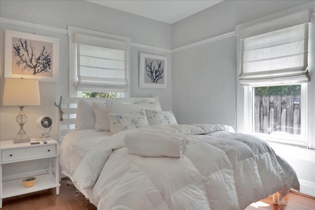A bright and serene bedroom features a spacious bed with white bedding and decorative pillows. Soft curtains filter natural light from the window, and framed artwork adorns the walls. A nightstand with a lamp and small decorative bowl is positioned beside the bed.