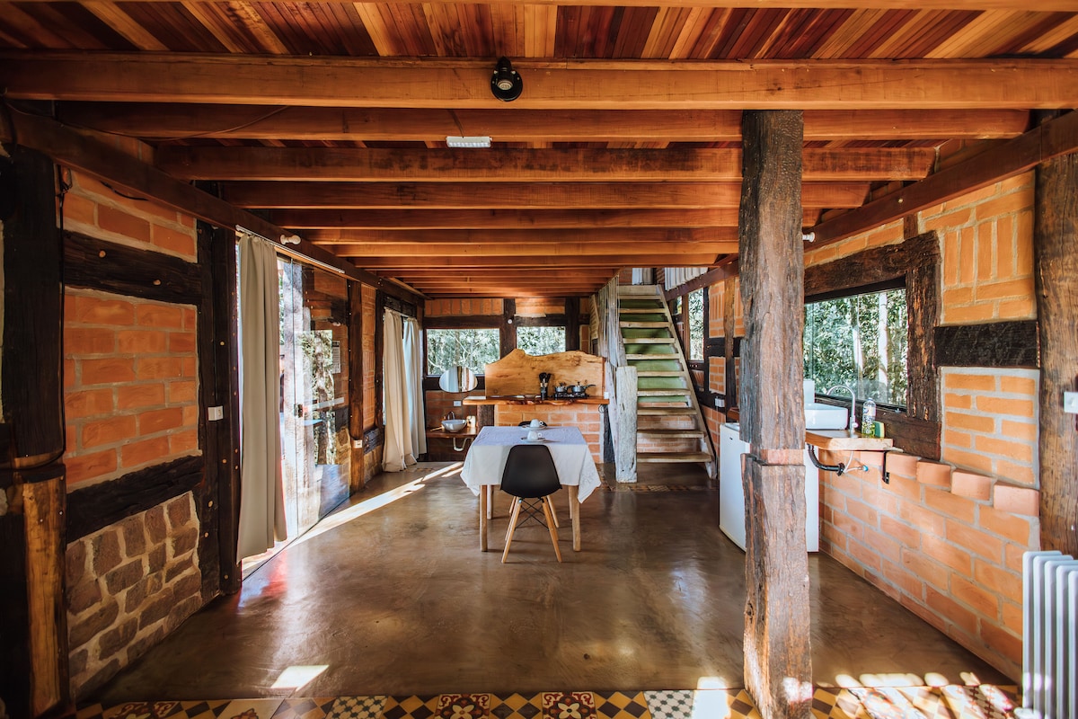 An open living area features natural wood beams and stone accents. A dining table is positioned centrally, surrounded by natural light filtering through large windows. A staircase leads to the upper level, while a kitchenette is set against the far wall, complementing the rustic decor.