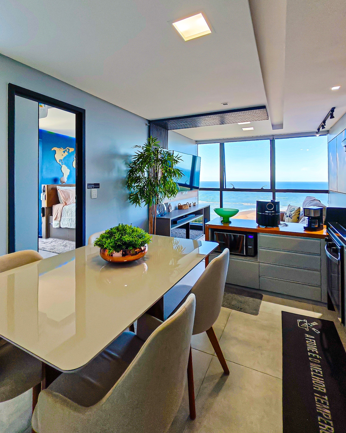 A modern dining area is enclosed by large windows, providing a panoramic view of the beach. The sleek table is surrounded by four chairs, while a vibrant plant centers the table. Kitchen appliances and a television are visible in the background.
