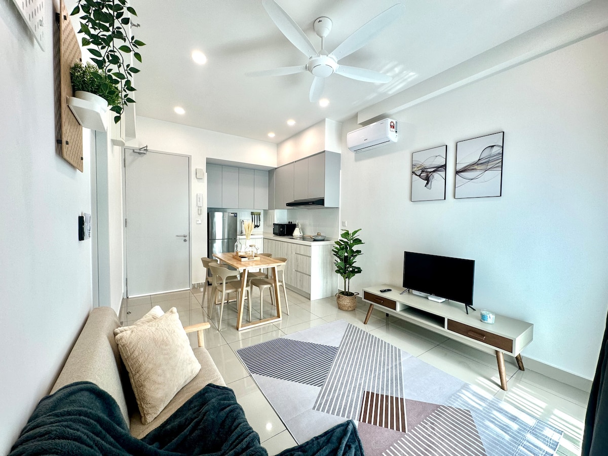 A minimalist living area is presented with a light-coloured sofa and a cozy blanket draped over it. A small dining table, equipped with two chairs, is visible. A television rests on a low console, and potted plants add greenery to the space.
