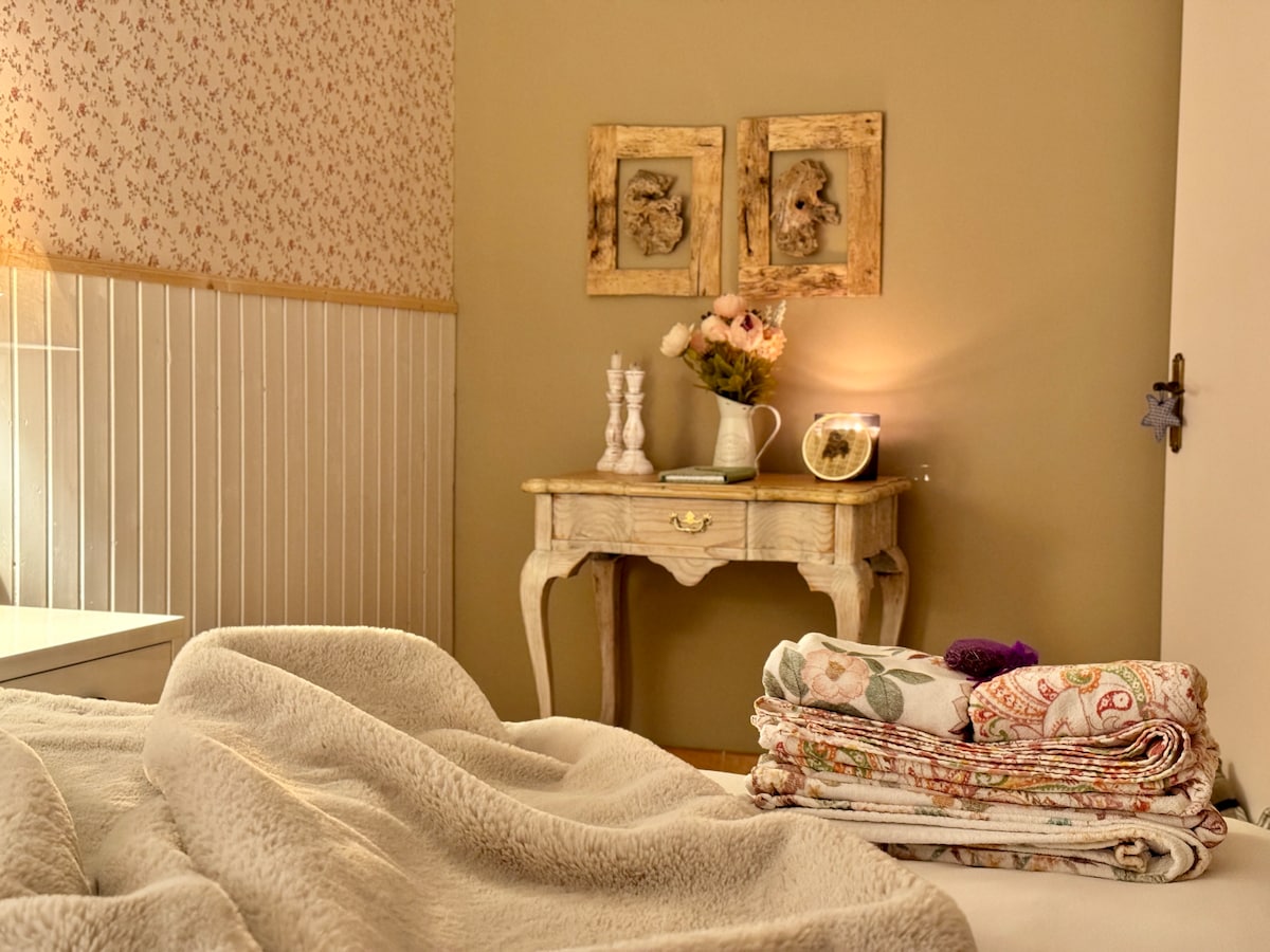 A tranquil bedroom setting features a neatly made bed with soft blankets. A small table is adorned with a vase of flowers and decorative frames on the wall. Natural light enhances the calming color palette of the decor.