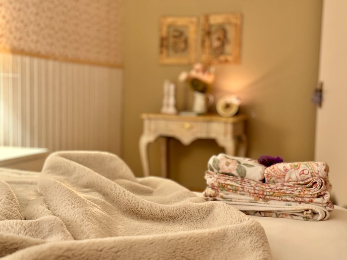 A neatly made bed is adorned with a soft, creamy throw blanket resting atop. In the background, a small decorative table holds a vase with fresh flowers and a framed artwork, contributing to a calm and welcoming atmosphere.