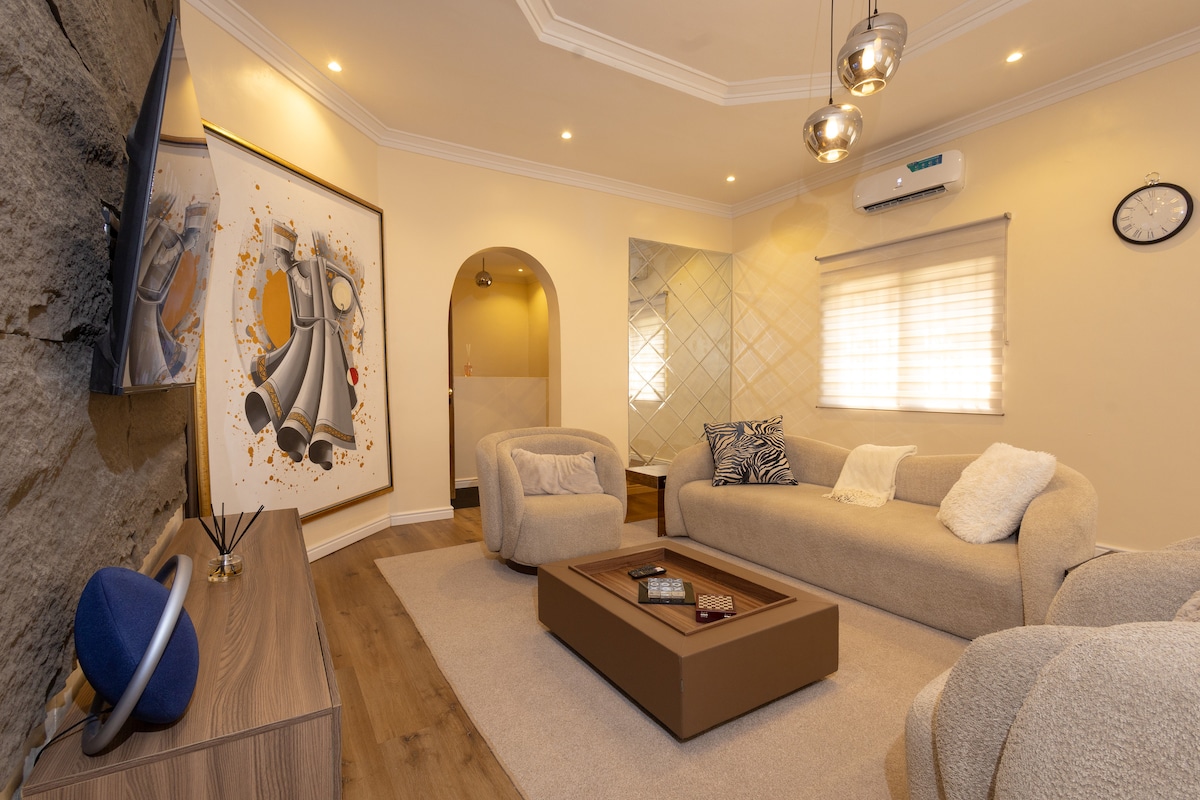 A welcoming living area features comfortable seating with soft, neutral tones. A large artwork adorns one wall, while a cozy coffee table occupies the center space. Ambient lighting is provided by stylish fixtures, and a wall-mounted TV complements the decor. The room is airy with natural light filtering through partially covered windows.