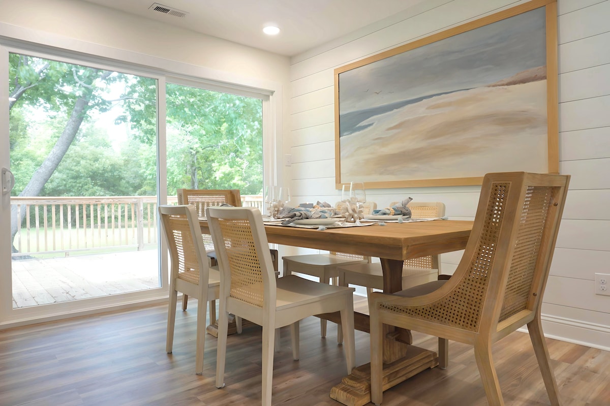 A dining area features a wooden table surrounded by six chairs, designed with a combination of open backs and cushioned seats. A large artwork depicting a serene coastal scene hangs on the wall. Natural light fills the space through sliding glass doors leading to a wooden deck.