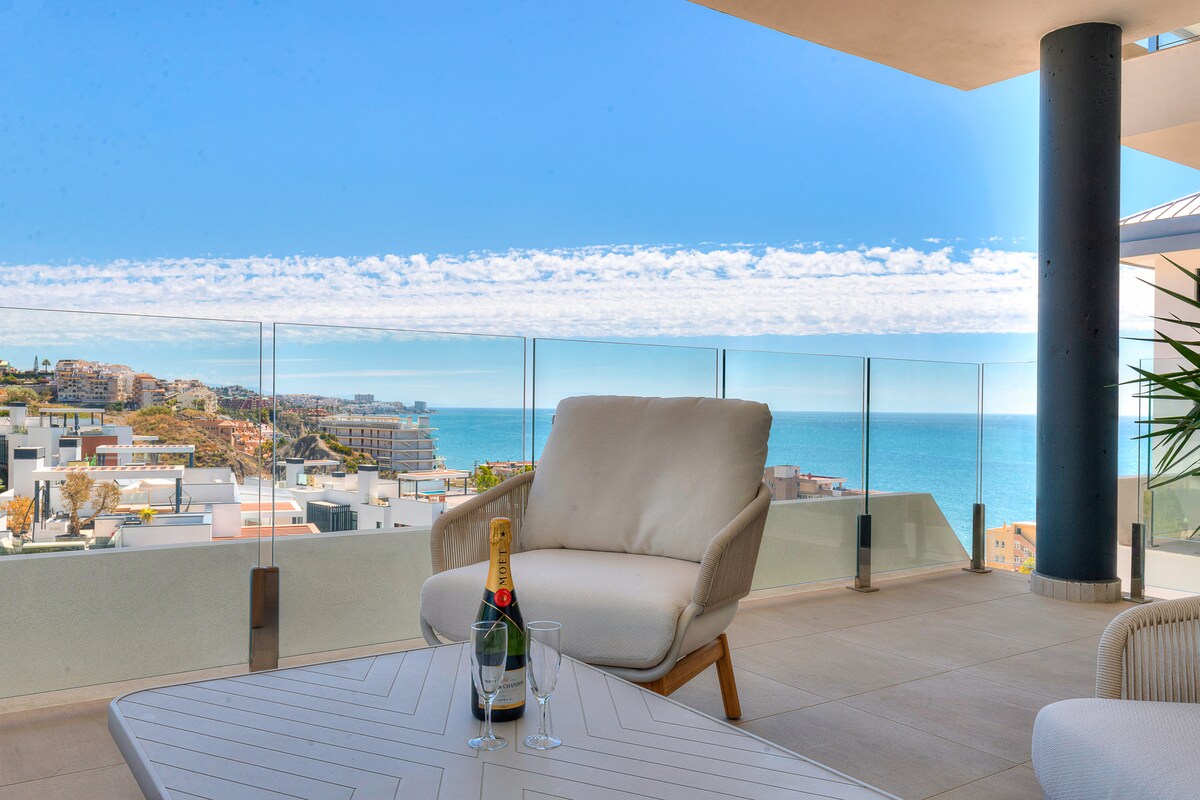 A spacious terrace features comfortable seating overlooking the ocean. A glass table holds a bottle of champagne and two glasses. The clear blue sky enhances the view of the coastline, creating a serene atmosphere for relaxation.