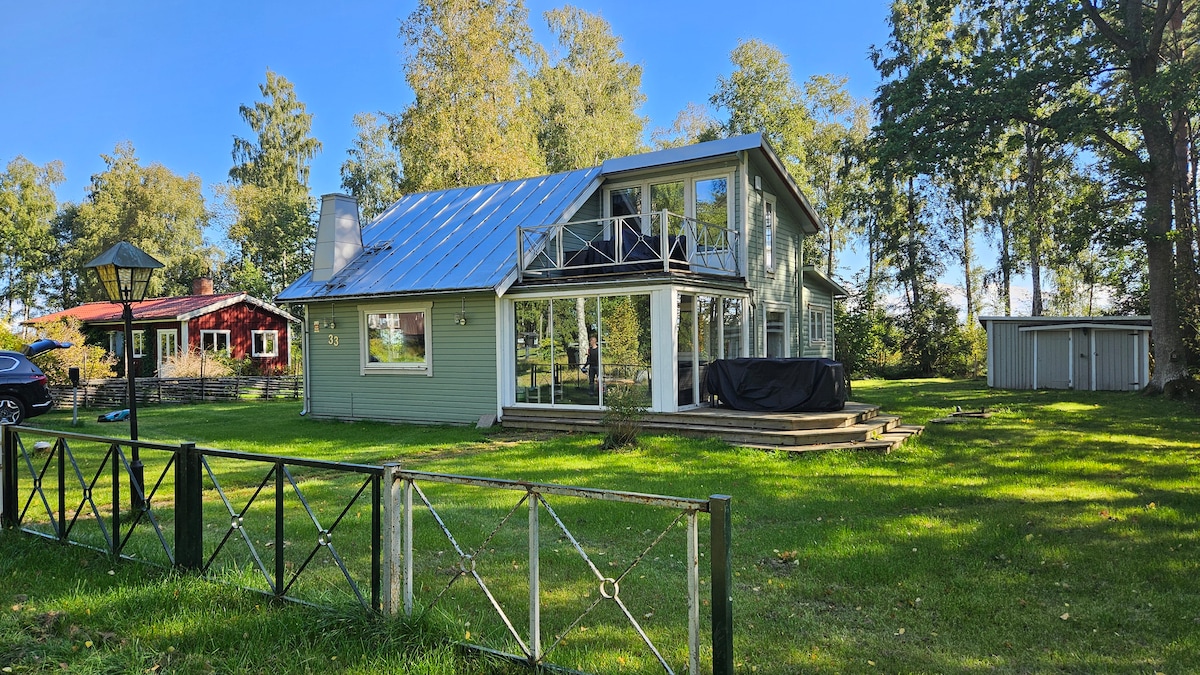 The exterior of the property features a green two-story house with a sloped roof and large glass doors that provide a view of the surrounding landscape. Lush grass covers the yard, accompanied by trees and nearby buildings, including a red structure and a shed.