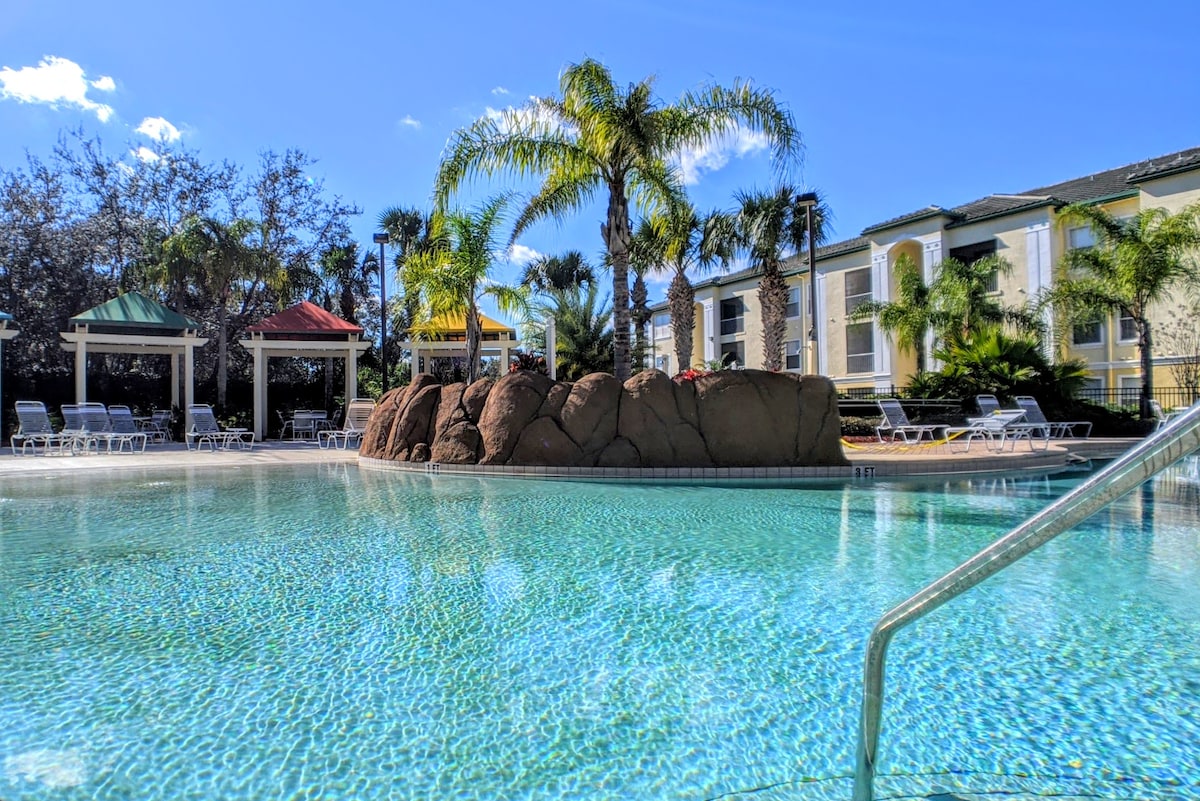 A serene swimming pool features a smooth, clear surface reflecting palm trees and sky. A rocky island is positioned in the center, surrounded by lush greenery. Lounge chairs are arranged around the pool area, under shaded gazebos, creating a relaxed outdoor space.