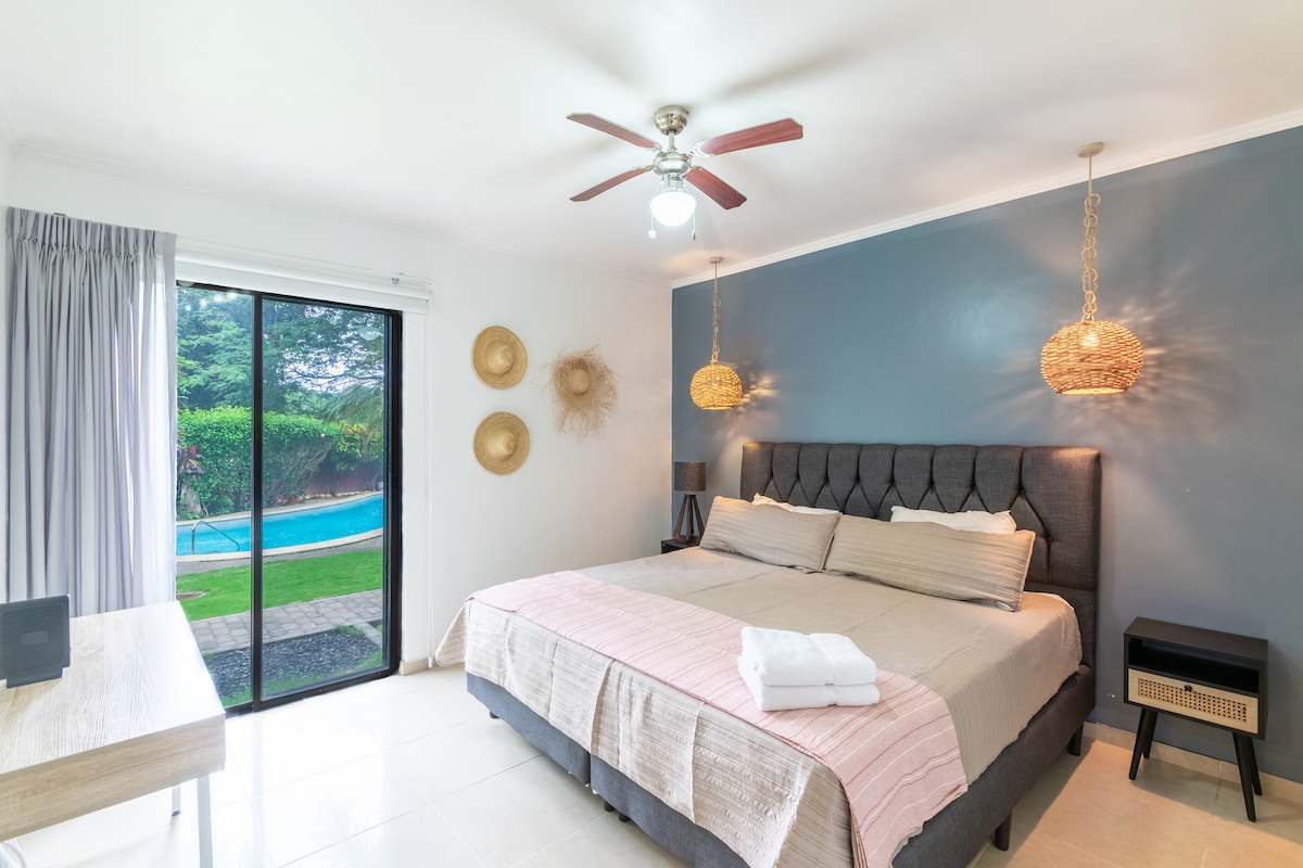 A spacious bedroom is shown, featuring a large bed with neutral bedding. Two woven pendant lights hang above the bed, complementing a modern design. A ceiling fan is visible, and large glass doors open to a green view of the poolside terrace, allowing natural light to enhance the space.