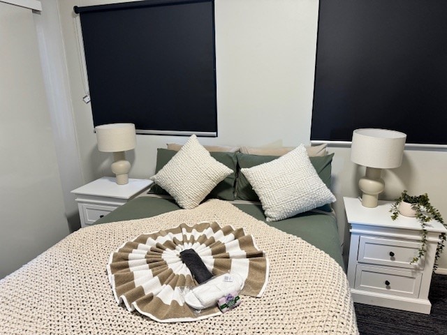 A serene sleeping area features a spacious bed adorned with a textured blanket in neutral tones. Two decorative pillows enhance the bedding. Nightstands with soft lamps flank the bed, and dark window coverings provide privacy in the room.