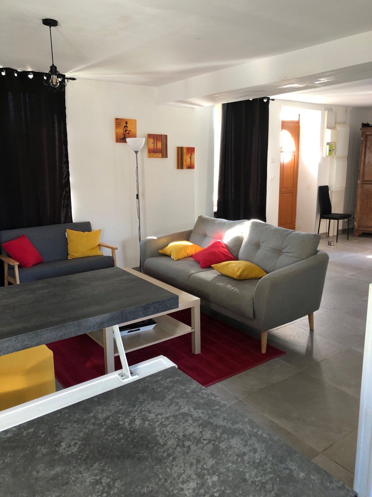 The living area features a combination of seating options, including a gray sofa and two colorful chairs, arranged around a coffee table. A vibrant rug adds warmth to the space, which is illuminated by natural light through large windows, creating a cozy atmosphere.