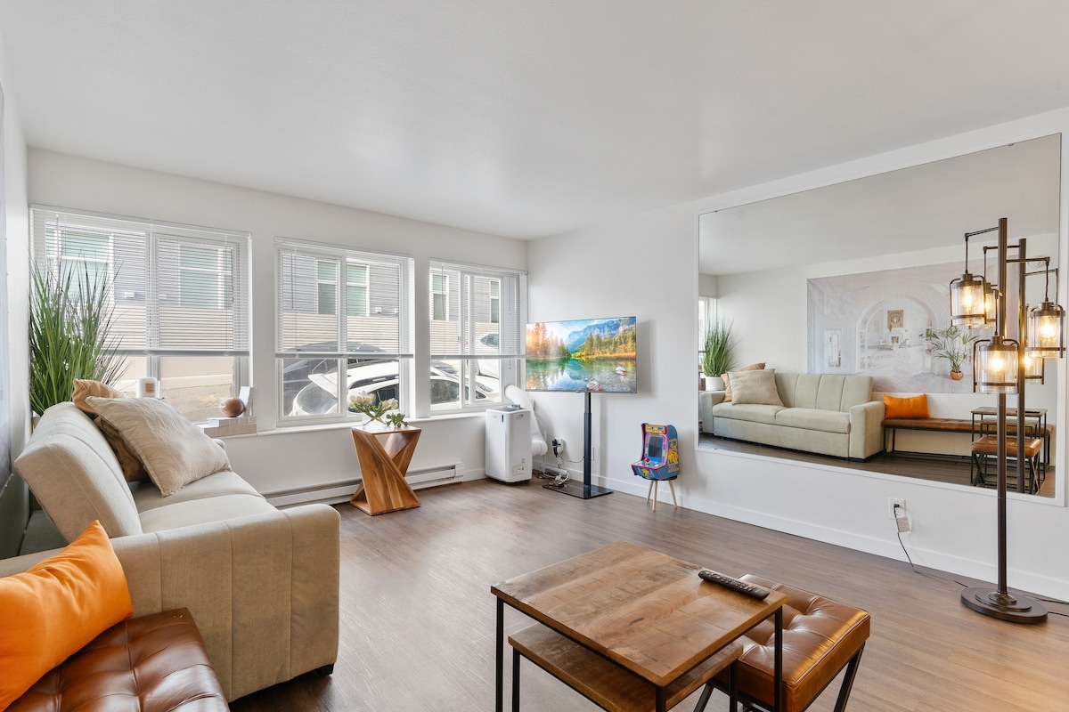 A spacious living area features large windows allowing natural light to fill the room. A comfortable couch is accompanied by a transitional coffee table and a stylish floor lamp. A wall-mounted smart TV displays vibrant artwork, while a large mirror enhances the sense of openness.