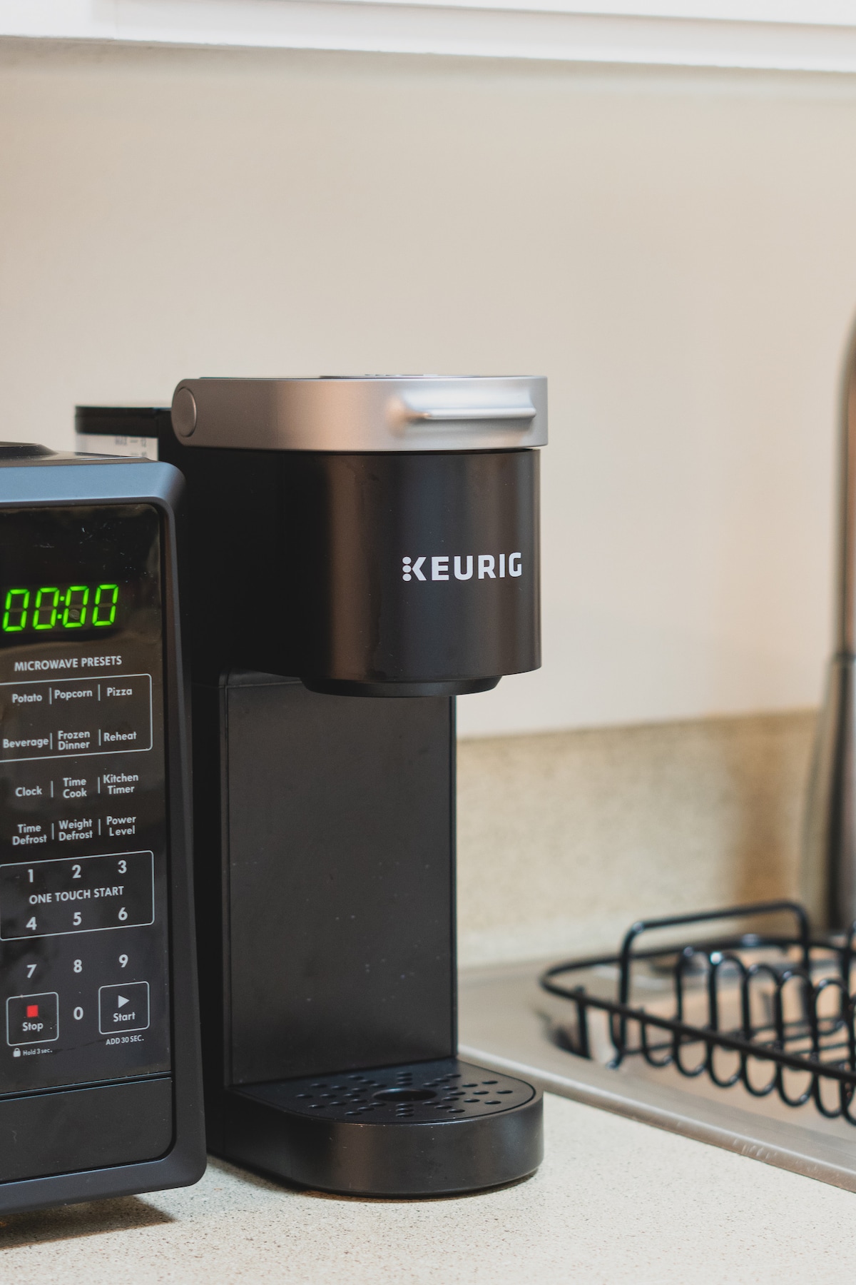 A modern coffee maker is seen on a kitchen countertop, featuring a sleek black design and a single-serve functionality. A microwave with digital controls is positioned beside it, indicating a well-equipped kitchen space for meal preparation and convenience.