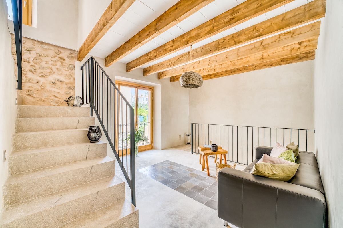 A spacious living area features exposed wooden beams overhead, with a staircase leading to the upper floor. Natural light enters through large windows, highlighting a simple seating arrangement with a coffee table. The flooring combines light stone with dark tiles, creating an inviting contrast.