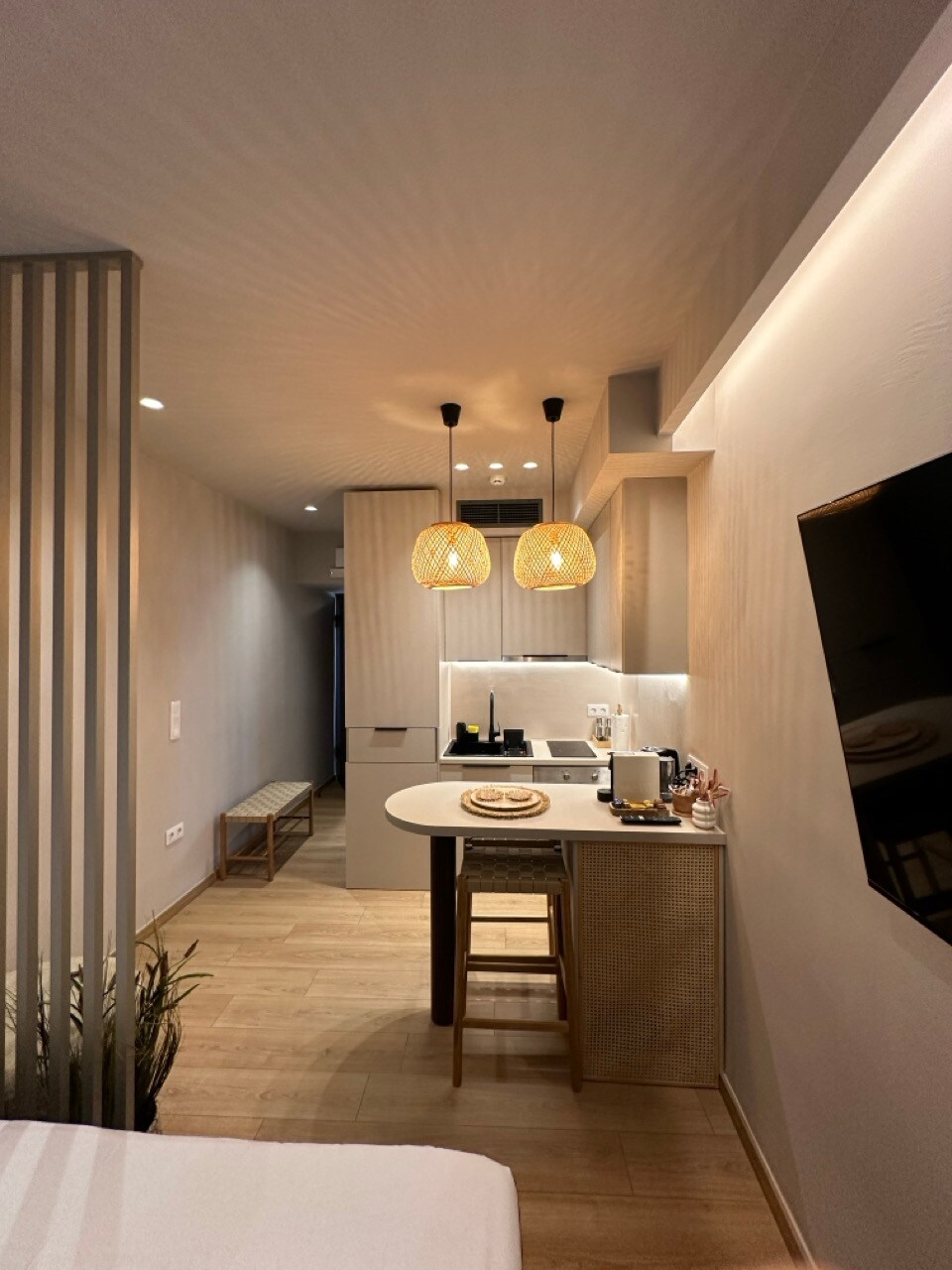 A modern kitchenette is visible, featuring light wood cabinetry and two pendant lights. A round table with two chairs sits nearby. The space is illuminated with soft lighting, creating a welcoming atmosphere. A television is mounted on the wall opposite the sleeping area.