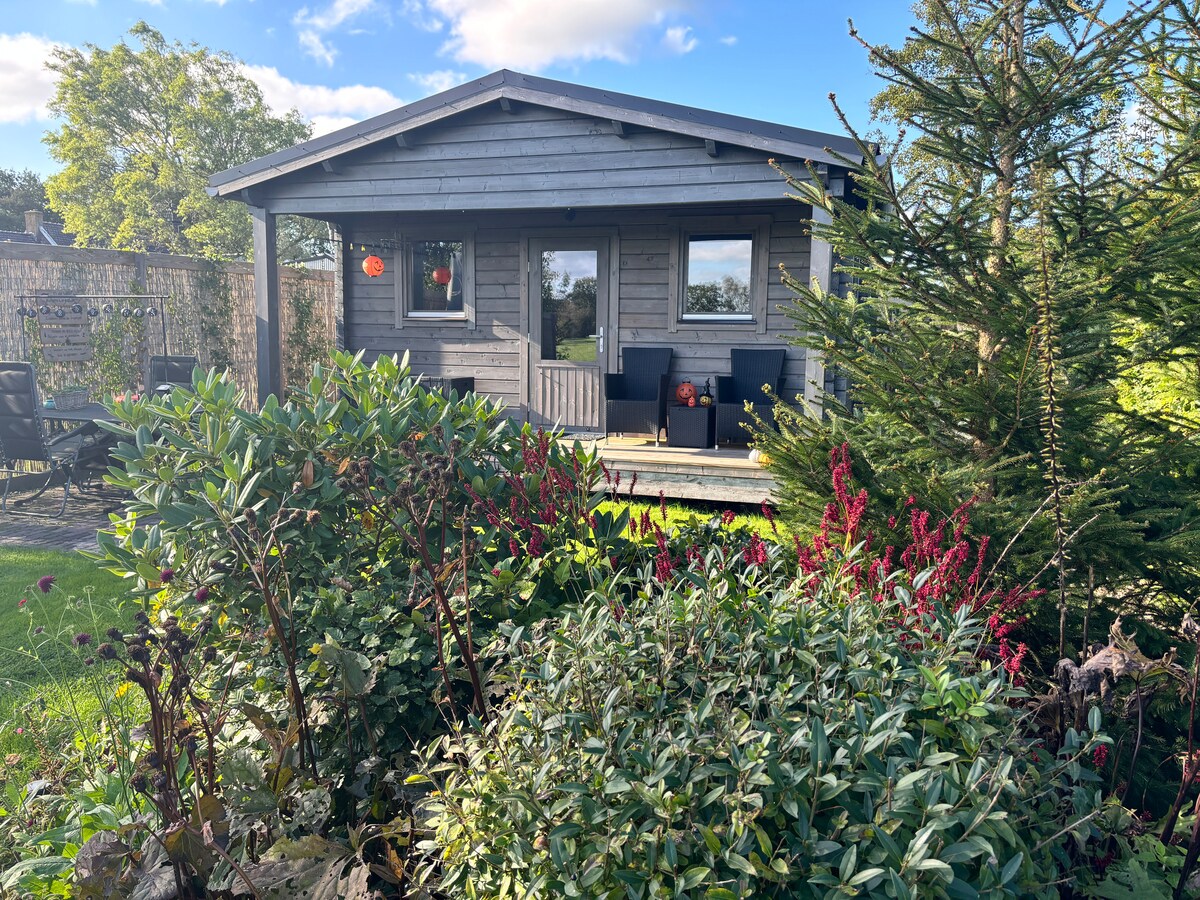 A charming wooden vacation house is surrounded by greenery and colorful flowers. The exterior features a porch with seating and large windows, allowing for natural light. A vibrant garden complements the serene setting, creating a peaceful atmosphere.