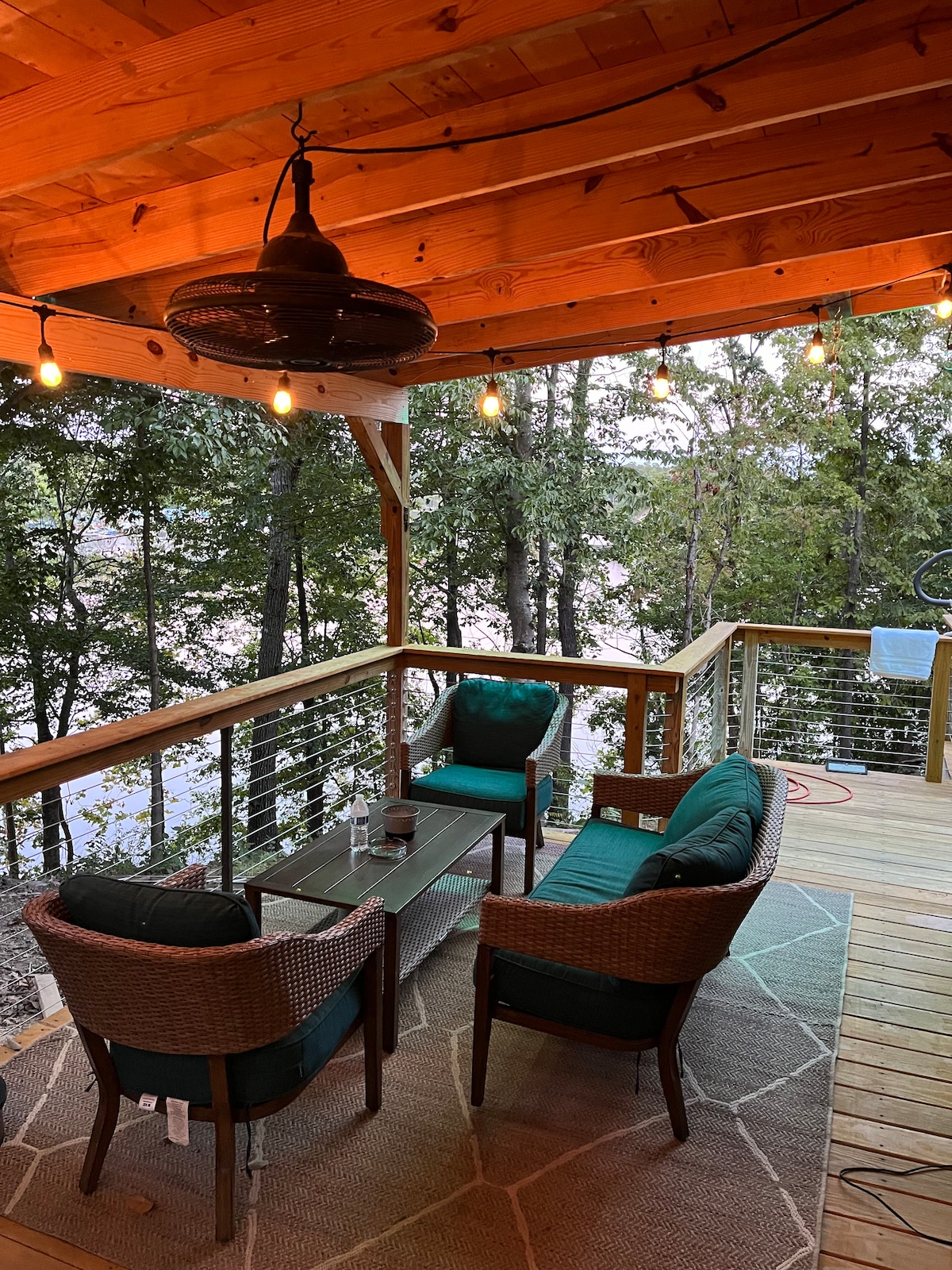 A covered deck is shown, featuring comfortable seating arrangements with green cushions. A small table is placed at the center, surrounded by a textured area rug. Soft lighting is provided by hanging bulb lights, enhancing the peaceful view of surrounding trees and the lake.