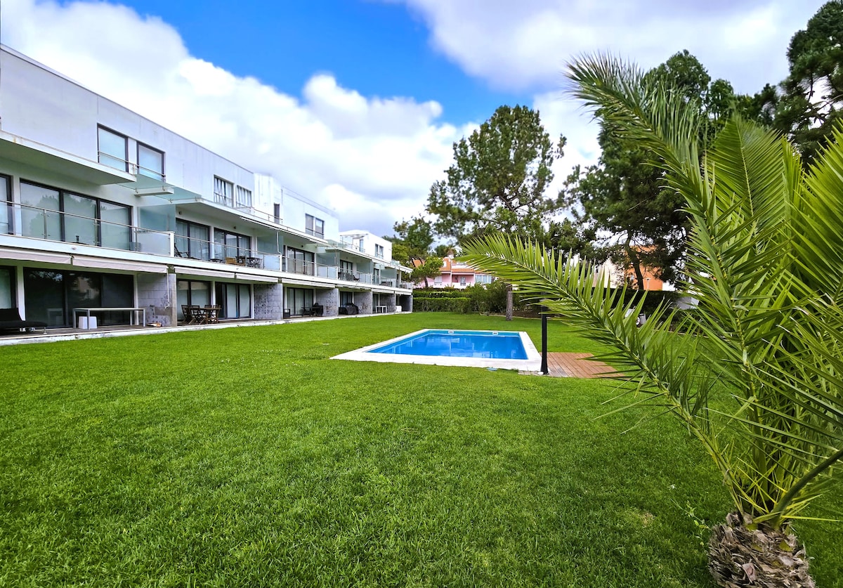 A well-maintained green lawn features a rectangular pool surrounded by a wooden deck. The two-story condominium buildings are visible in the background, each with balconies overlooking the landscaped grounds. Lush palm trees add a tropical touch to the serene outdoor space.