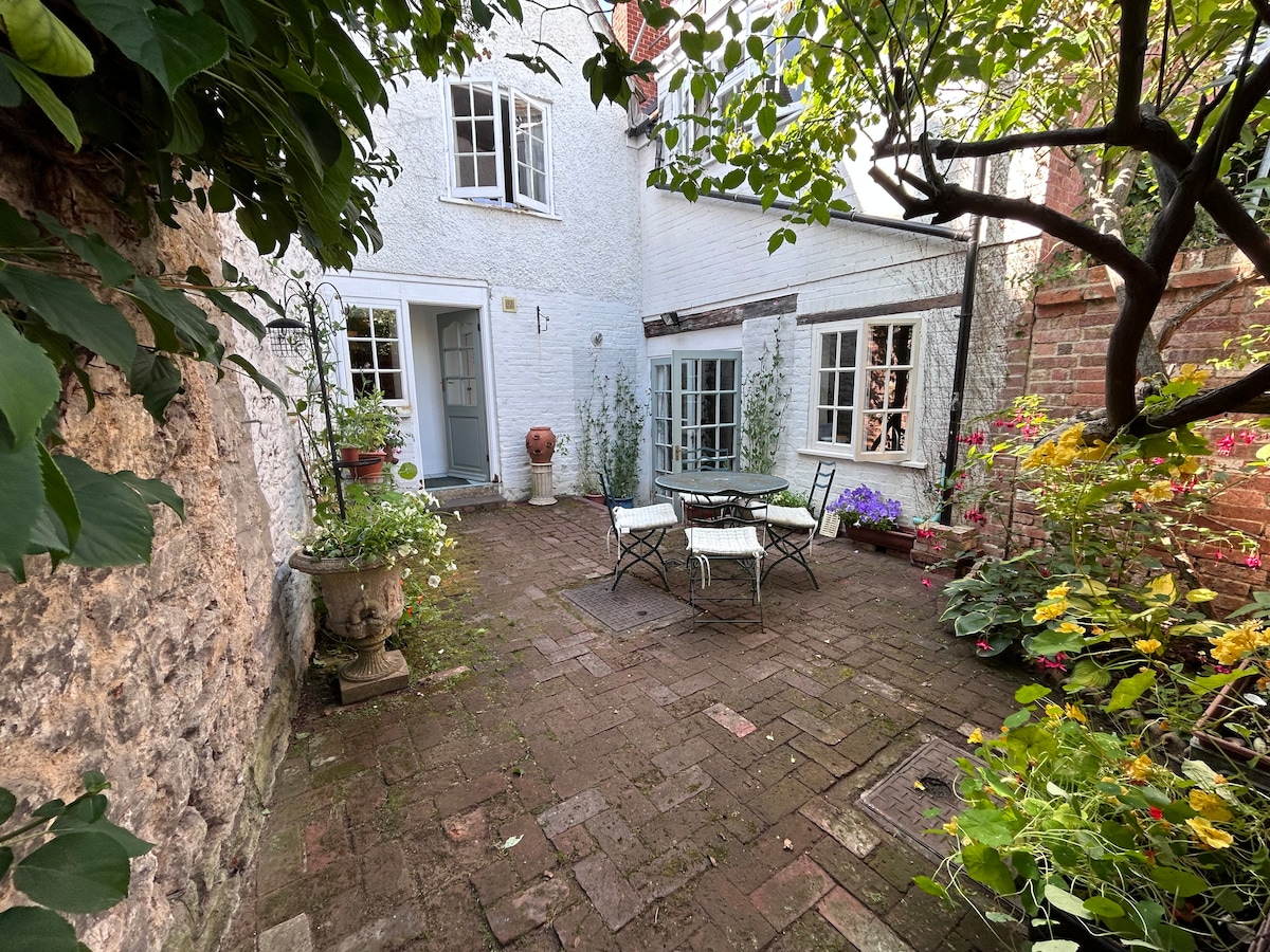 A small patio garden features a stone-paved area with a table and chairs for outdoor dining. Surrounding plants in various colors provide a lush and inviting environment. White cottage walls and large windows create a charming backdrop for this serene space.