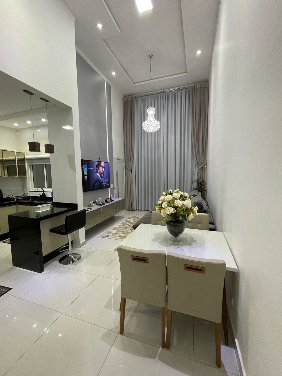A spacious common area features a modern dining table with light-colored chairs, complemented by a decorative vase of flowers. A large television is mounted on the wall, with soft lighting provided by an elegant chandelier above. Sleek cabinetry and a glimpse of the kitchen can be seen in the background.