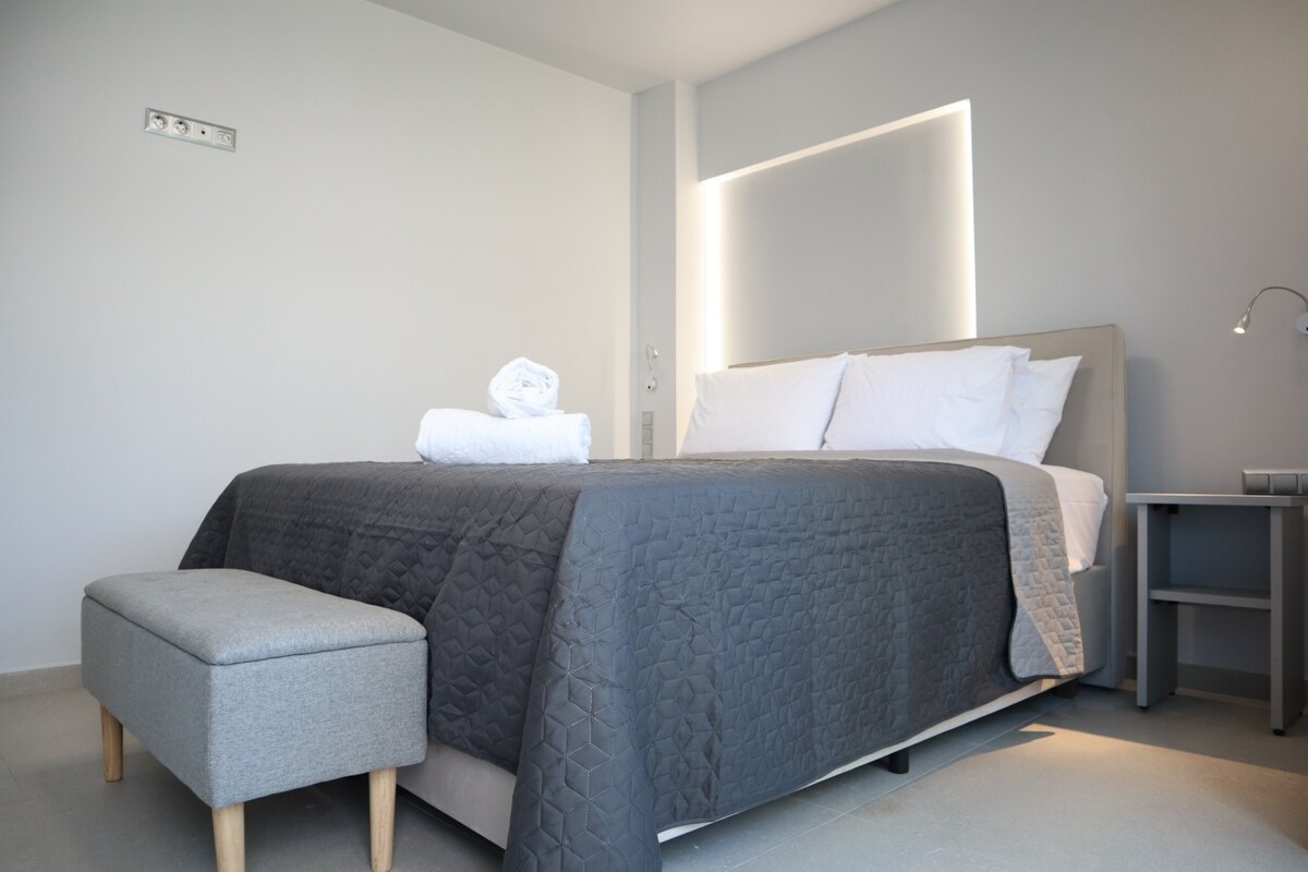 A well-appointed sleeping area features a spacious bed with neatly arranged white linens and a textured gray coverlet. A comfortable bench sits at the foot of the bed, complemented by a bedside table with a lamp against the subtle gray wall.