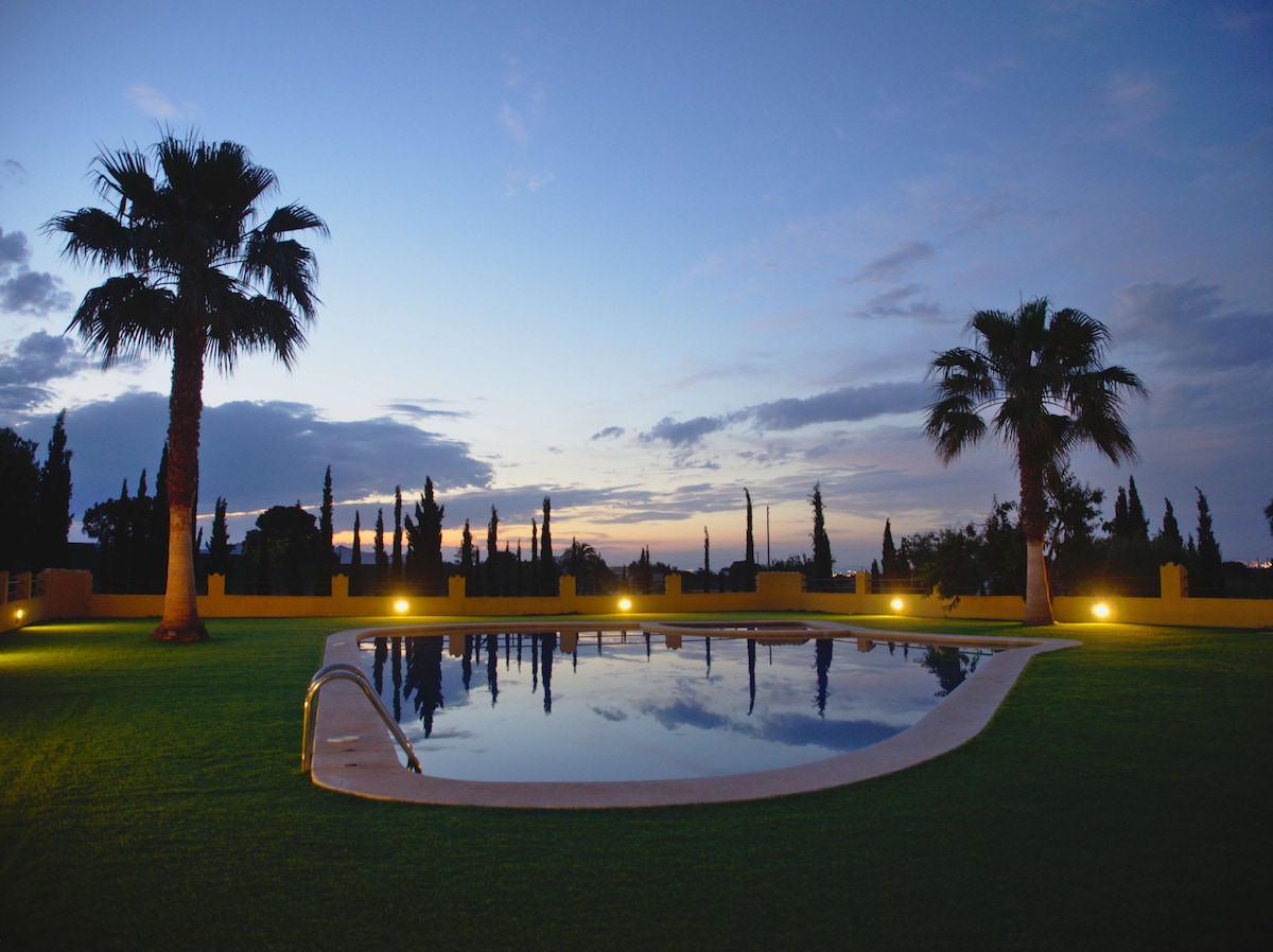 A tranquil swimming pool is set against a twilight sky, surrounded by lush green grass. Palm trees frame the scene, and soft lights illuminate the area, creating a serene atmosphere ideal for relaxation.