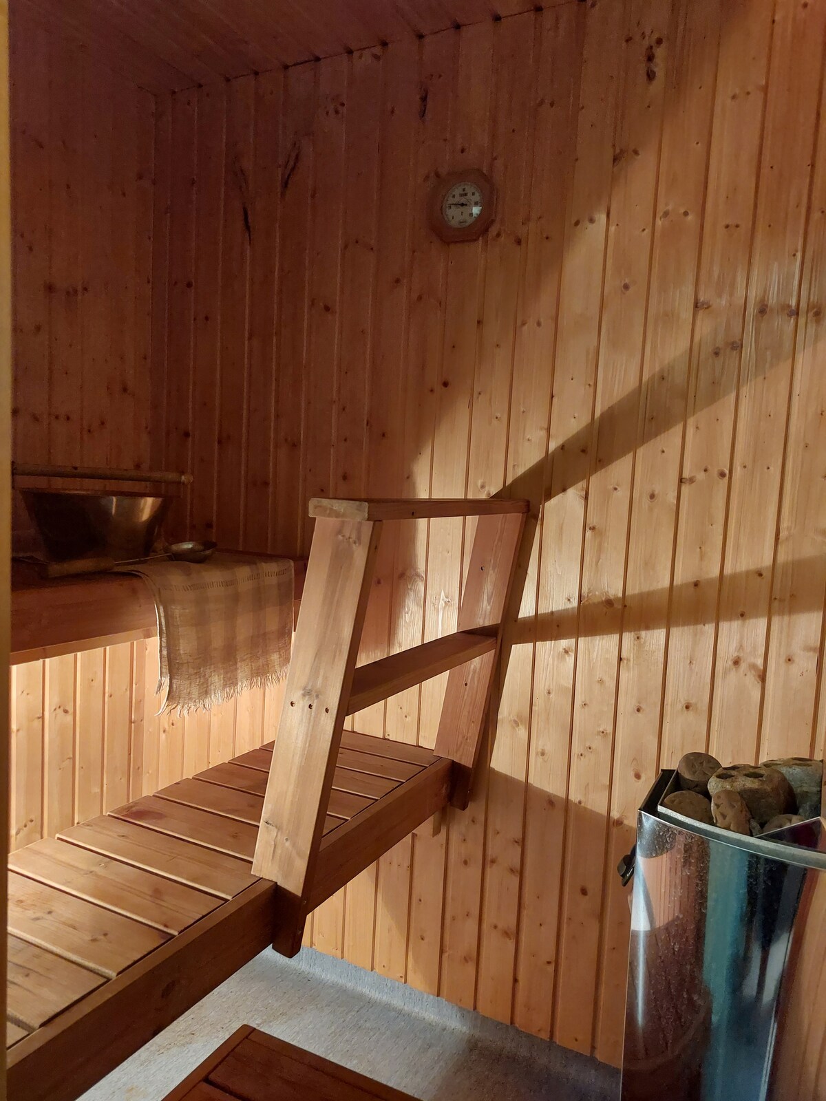 A wooden sauna features warm-toned planked walls and seating. A small clock is mounted on the wall, indicating the time. A metal bucket and ladle rest on a shelf, while a storage container holds firewood, contributing to the sauna's functional ambiance.