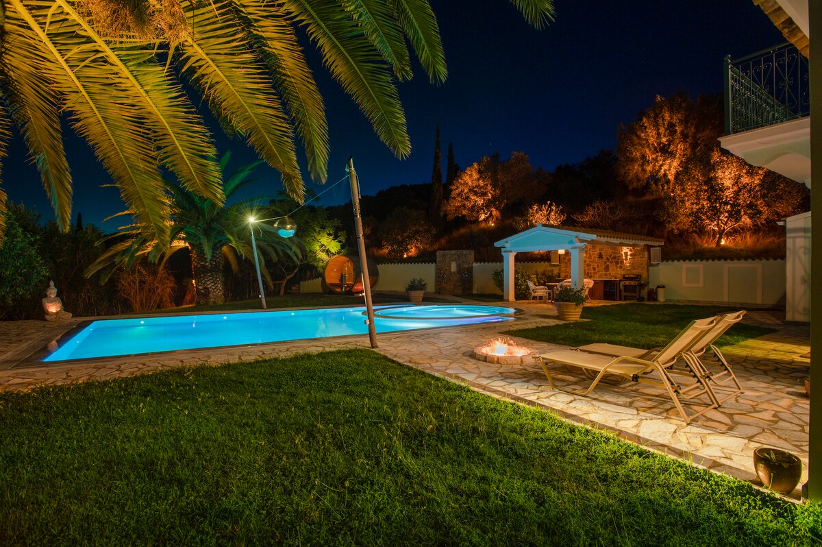 The outdoor area is illuminated at night, featuring a heated pool surrounded by lush greenery. Lounge chairs are positioned nearby, with soft lighting highlighting the palm trees and surrounding plants, creating a serene and inviting atmosphere.