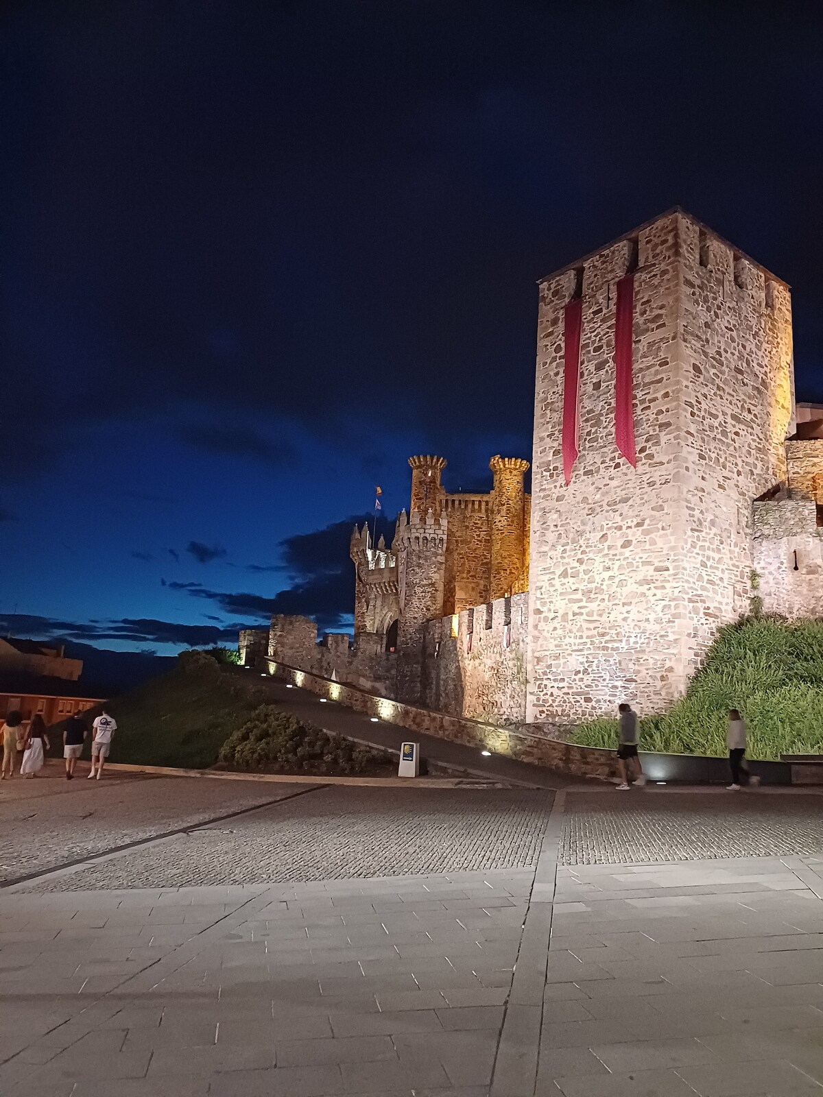 A medieval castle tower is illuminated by warm light against a deep blue evening sky. The stone walls feature banners hanging from the tall structure, while the surrounding landscape includes pathways and green areas, creating a tranquil night scene.