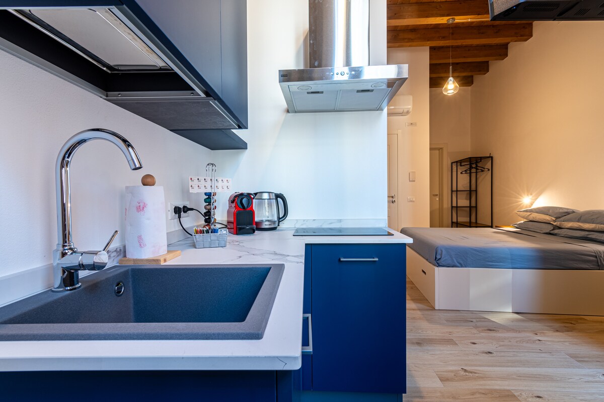A modern kitchenette is showcased with dark blue cabinets and a sleek white countertop. A stainless steel sink and hood are visible above the stove. In the background, a cozy sleeping area features a large bed with grey linens, illuminated by natural light from the adjacent window.