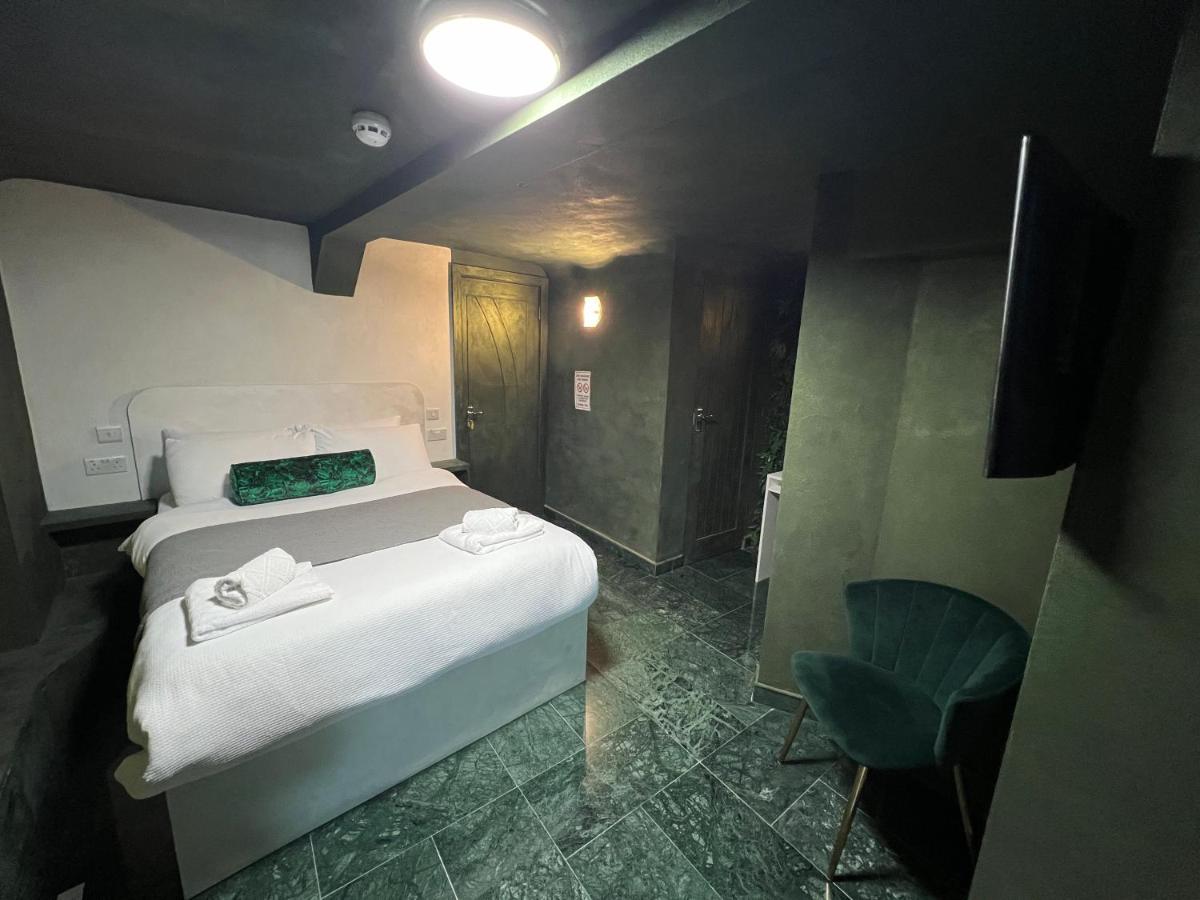 A modern guest room features a spacious bed with neatly arranged linens. A green accent pillow adds a touch of color. Dark stone flooring complements the sleek design, while a small green chair sits next to the bed. A flat-screen TV is mounted on the wall.