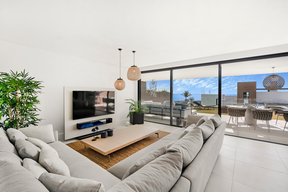 A spacious living room is designed with large windows that allow natural light to fill the space. A comfortable sectional sofa faces a modern entertainment unit, accompanied by a stylish coffee table. The area features indoor plants, enhancing the connection with nature.