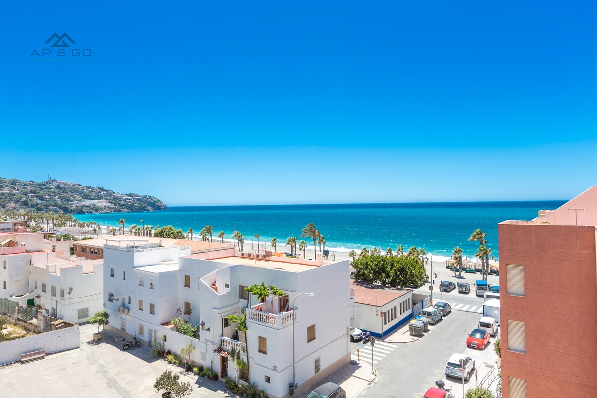 A panoramic view showcases the shimmering coastline with the azure sea stretching towards the horizon. In the foreground, modern buildings are positioned along a clean street, palm trees providing a touch of greenery against the clear blue sky.