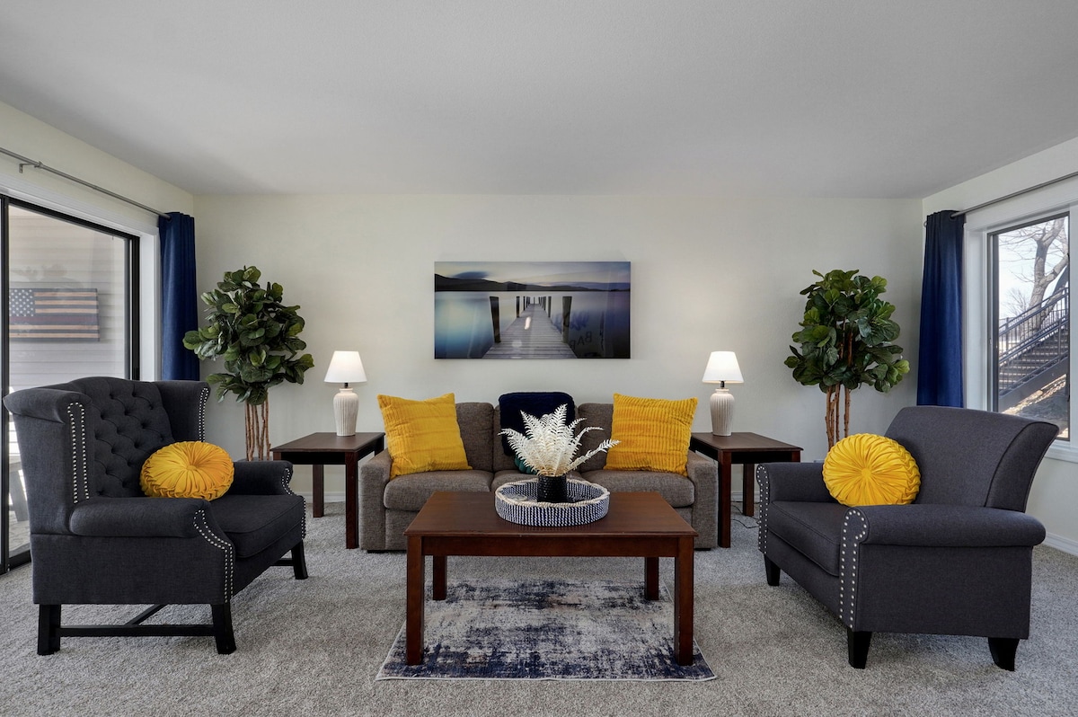 The living area offers comfortable seating with two armchairs and a sofa, accented by yellow cushions. A wooden coffee table is centered in the space, with a decorative arrangement on top. Tall plants flank the seating, and light filters in through large windows on either side.