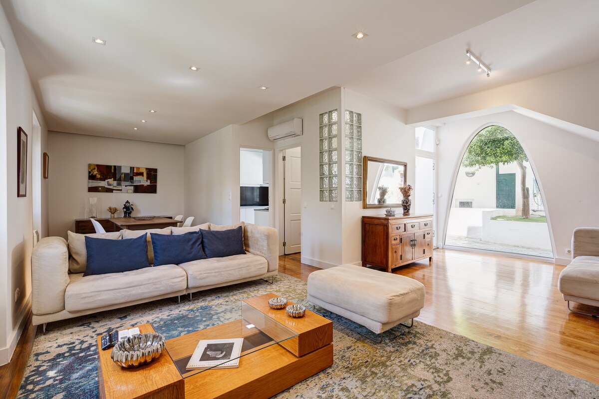 The spacious living area is highlighted by a soft, neutral sofa adorned with blue cushions. A wooden coffee table is centrally placed on a patterned rug, while light fills the room through large windows, creating a bright and welcoming ambiance.