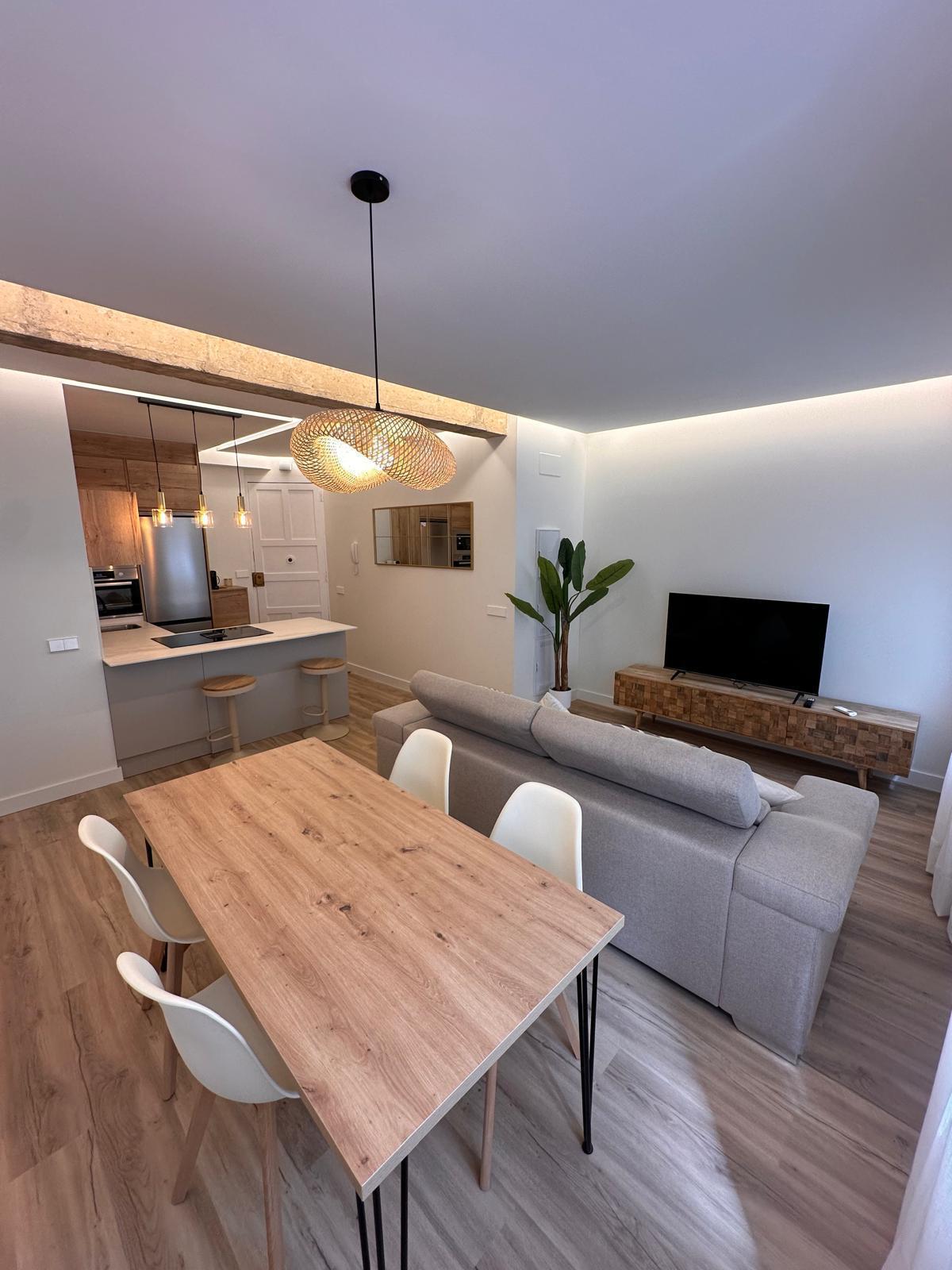 A modern dining area features a wooden table paired with four minimalist chairs. A comfortable sofa faces a wood-accented media console with a television. Pendant lighting adds warmth, while a small plant introduces a touch of greenery to the space.